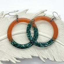 Large resin hoop earrings with a split-tone design featuring burnt orange shimmer and crushed blue green glitter roxs. These handmade statement earrings are displayed on a white feather, showcasing the translucent autumn colors and sparkling texture. Perfect for boho style enthusiasts or as a unique jewelry gift, these lightweight circular dangles offer a bold, colorful aesthetic for everyday wear or special occasions.