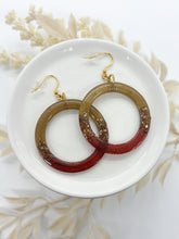 A pair of hoop earrings with gold-colored fishhook ear wires. The hoops transition from a golden-brown to a deep brown, with embedded glitter roxs. The earrings rest on a white plate.