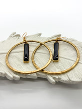 A pair of hammered statement hoop earrings displayed on a white ceramic feather. Each earring features a vertical, rectangular black resin bar suspended in the center of the hoop. The shot is taken at a slight angle, highlighting the texture of the metal and the glossy reflection on the black charms against a clean, minimalist white background.