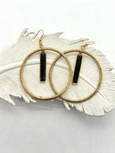 A pair of gold-toned hoop earrings featuring a weathered, hammered texture. Each hoop centers a vertical, rectangular black resin bar charm. The earrings rest on a white, decorative ceramic feather against a clean white background. The design blends a modern geometric aesthetic with vintage-inspired metallic finishes.