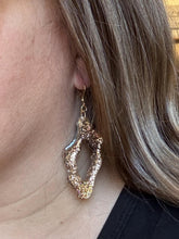 A close-up photograph of a woman with long blonde hair, seen from the side profile, wearing a single large, organic-shaped dangle earring. The earring is a neutral ombre sculptural piece with a sparkling or glittery texture, connected to a delicate gold french ear wire. It has an open, abstract loop design and hangs gracefully just below her earlobe. She is wearing a simple black top, and the background is a soft, natural indoor light.
