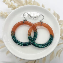 A pair of large resin hoop earrings displayed on a white ceramic dish, featuring a striking burnt orange and turquoise color palette. These handmade statement earrings combine a shimmering translucent top with a bottom half of crushed glass glitter for a textured, sparkling look. Designed with hypoallergenic hooks, these lightweight circular dangles are perfect for boho style outfits, autumn fashion, or a unique handcrafted jewelry