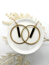 A pair of everyday boho hoop earrings resting on a white ceramic dish. Each large hoop features a dangling, vertical rectangular bar charm with a black resin inlay. The background is a crisp white, accented by soft, cream-colored dried foliage that adds a rustic, bohemian touch. The lighting is bright and clean, highlighting the weathered texture of the metal and the glossy finish of the black charms.