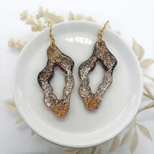 Handmade organic wavy resin earrings featuring a shimmering brown and champagne glitter gradient. These unique earth-tone dangles have a lightweight cutout design and hang from gold ear wires. Displayed on a white ceramic dish, they offer a boho-chic aesthetic perfect for neutral styling or a sparkly statement gift for her.