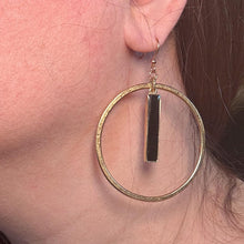 Close-up modeled photo of a gold hammered hoop earring worn on an ear. The earring features a large, textured open circle frame with a sleek vertical black resin-filled bar charm suspended in the center, hanging from a gold ear wire hook against a soft neutral background.