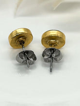 A close-up product photograph displaying the backs of a pair of handmade, rustic gold-plated bezel stud earrings. They rest face-down on a clean white surface, showcasing the detailed gold-plated finish on the round settings and the silver-toned, hypoallergenic surgical steel post and butterfly-back closures.