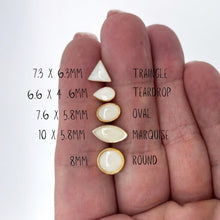 A size comparison of various white resin stud earrings held against a hand for scale. The image features six different geometric shapes in 24K gold plated settings, including triangle (7.3 x 6.mm), teardrop (6.6 x 4.6mm), oval (7.6 x 5.8mm), marquise (10 x 5.8mm), and a round size (8mm). These minimalist geometric studs are perfect for second piercings or everyday wear. Handmade jewelry size guide for scale and variety.