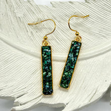 Handmade vertical rectangle bar dangle earrings displayed on a white textured ceramic feather background. The earrings feature a textured, mosaic-like inlay of crushed emerald green, dark teal, and shimmering blue green glass like fragments. The bars are framed in a sleek 24K gold plated bezel trim and hang from matching gold-plated hypoallergenic French hook ear wires equipped with clear rubber safety stoppers.