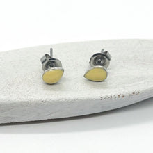 A pair of small, handmade teardrop stud earrings resting on a rustic, white-painted wooden surface against a clean white background. The earrings feature a delicate .999 fine silver-plated textured bezel filled with a smooth, pale yellow resin inlay. The hypoallergenic posts and butterfly backings are clearly visible, highlighting the modern-vintage, artisan design of the jewelry pieces.
