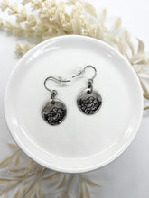 A top-down view shows a pair of rustic, fine silver-plated dangle earrings displayed on a small white ceramic dish, set against a blurred cream floral background. The earrings feature small, irregularly shaped silver discs, each ink stamped with an intricate, dark rose botanical design. They hang from curved stainless steel ear wires, which are equipped with small, clear rubber backings. They are sealed in a glossy resin for long lasting wear.