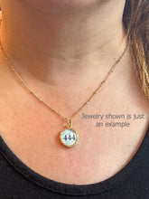 A close-up photograph shows a person wearing a delicate gold satellite ball chain necklace against their collarbone. The necklace features a small, round gold-rimmed pendant with a light blue watercolor background displaying the angel number "444" in a clean black font. The person is wearing a casual black scoop-neck top. Jewelry shown is just an example.