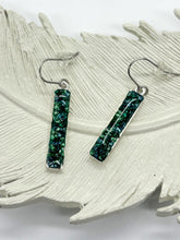 Handmade vertical rectangle bar dangle earrings displayed on a white textured ceramic feather background. The earrings feature a textured, mosaic-like resin inlay of crushed emerald green, deep teal, and shimmering dark stone fragments. The bars are framed in a sleek .999 fine silver plated bezel trim and hang from matching silver-colored hypoallergenic French hook ear wires equipped with clear rubber safety stoppers.