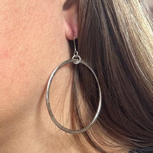 A close-up side view shows a woman with long brown hair wearing a large, lightweight hammered silver hoop earring. The large open-circle hoop has an artisan, textured finish with a subtle vintage patina. It hangs elegantly from a small, decorative rope-textured connector ring attached to a thin, hypoallergenic surgical steel wire hook, resting flat against the cheek to showcase its bohemian style.