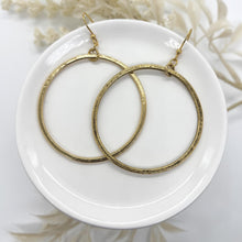 A pair of large, minimalist gold hoop earrings resting on a small, glossy white ceramic dish. The hoops have a thin, flat profile and features a rustic, hammered metallic texture. They hang from delicate gold fishhook ear wires attached to the top of each hoop. In the soft-focus background, cream-colored dried floral sprigs border the top and bottom corners of the frame against a clean, bright white backdrop.