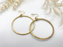 A pair of large, thin gold hoop earrings lies flat on a bright white surface, overlapping slightly at the center. The thin hoops feature a flat profile with a textured, hammered metallic finish. They hang from gold fishhook ear wires fitted with clear plastic backings. The background includes out-of-focus decorative elements: a textured white branch in the upper-left corner and soft, cream-colored dried leaves framing the upper-right side.