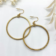 A pair of large, hammered gold hoop earrings displayed on a small, glossy white ceramic dish. The hoops feature a modern-vintage look with flat, thin edges and a textured, slightly hammered metallic surface. They hang from delicate gold fishhook ear wires attached to the tops of the hoops. Out-of-focus, creamy-white dried floral sprigs frame the dish from the top and bottom corners, set against a clean, bright white background.