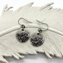 A pair of handcrafted, rustic bohemian dangle earrings displayed on a textured white decorative ceramic feather. The earrings feature round, fine silver-plated disc charms with an ink stamped black-lined rose flower design. The charms hang from hypoallergenic surgical steel ear wires equipped with clear rubber backings.