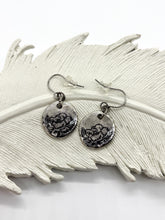A pair of handcrafted, rustic bohemian dangle earrings displayed on a textured white decorative ceramic feather. The earrings feature round, fine silver-plated disc charms with an ink stamped black-lined rose flower design. The charms hang from hypoallergenic surgical steel ear wires equipped with clear rubber backings.
