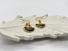 An angled, eye-level close-up shot shows a pair of rustic, gold-plated dangle earrings resting on a textured white ceramic feather dish. The perspective highlights the organic, uneven surfaces of the round coin charms. The earrings are positioned with one charm slightly overlapping the feather's center ridge, displaying the matching gold-toned fishhook ear wires and a clear rubber backing against a crisp white background.