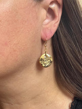 A close-up side profile shows a woman wearing a handcrafted gold-plated dangle earring. The earring features a small, rustic round disc charm with a dark, etched botanical rose design. The gold coin charm hangs from a thin, matching gold-toned fishhook ear wire. Her long brown hair is tucked behind her ear to cleanly display the lightweight, vintage-style jewelry against her skin.