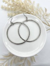 A top-down product photograph displays a pair of large hammered silver hoop earrings beautifully arranged on a glossy white ceramic dish. The open-circle hoops showcase an organic, hand-forged texture and are connected to thin French hook ear wires by small, twisted rope-style rings. Soft, cream-colored faux botanicals decorate the white background, highlighting a clean, modern-vintage bohemian aesthetic for artisan jewelry.