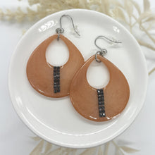 A pair of handmade peach-colored resin teardrop dangle earrings featuring an open cutout center. Each earring has a vertical, shimmering stripe of tiny black rhinestones accented near the bottom rim. They hang from silver fishhook ear wires with clear plastic backs, displayed flat on a glossy white ceramic round plate against a soft background with out-of-focus dried white flowers.