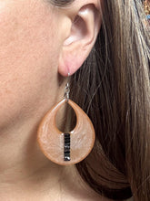 A close-up shot showing a person with long brown hair wearing one of the peach-colored resin teardrop earrings to demonstrate how it hangs and its relative size. The large, lightweight statement earring features an open cutout teardrop shape with a vertical stripe of sparkling black rhinestones. It hangs comfortably from a silver fishhook ear wire on the model's earlobe.