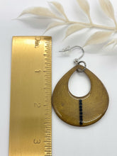 A large, handmade teardrop dangle earring in olive green shimmer resin with a vertical stripe of tiny black rhinestones. The open cutout earring rests flat next to a brass measuring ruler on a white background to show scale, measuring just over 2.5 inches long from the fishhook wire to the bottom edge. Text at the bottom reads, "Jewelry shown is an example of shape & size," with a soft sprig of pale dried leaves visible at the top.