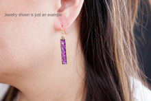 A close-up profile view of a person wearing a dangle earring. The earring features a vertical rectangular bar pendant with a thin gold border, filled with textured, glittering purple crushed stones. The pendant hangs from a thin gold wire hook. Text in the upper left corner reads, "Jewelry shown is just an example."