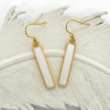 A pair of dangle earrings featuring vertical, rectangular bar pendants with a glossy white resin inlay and thin gold borders. The bars hang from gold fishhook ear wires. The earrings are displayed on a white, textured ceramic or plaster decorative feather dish against a plain white background.