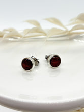 A close-up shot of a pair of handmade round stud earrings resting on a white ceramic dish. The minimalist jewelry features a smooth silver bezel filled with a solid dark brown shimmering resin inlay. One earring faces forward, while the other is slightly angled to show its side profile and surgical steel post hardware. A soft, out-of-focus white leaf branch decorates the background.