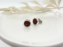 Close-up of a pair of handmade round stud earrings resting on a white ceramic dish. The minimalist jewelry features a textured silver bezel filled with a dark chocolate brown shimmering glitter resin inlay. One earring faces forward, while the other is angled to show its side profile and surgical steel post. A soft, out-of-focus white leaf branch is visible in the background.