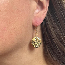 A close-up side profile shows a woman wearing a handcrafted gold-plated dangle earring. The earring features a small, rustic round disc charm with a dark, etched botanical rose design. The gold coin charm hangs from a thin, matching gold-toned fishhook ear wire. Her long brown hair is tucked behind her ear to cleanly display the lightweight, vintage-style jewelry against her skin.