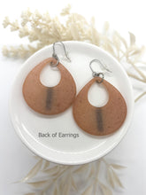The back of a pair of handmade peach-colored resin teardrop dangle earrings. The smooth, matte reverse side reveals a shadowed, dark vertical stripe where the front rhinestones are attached. They are shown lying flat on a round white ceramic dish with fishhook ear wires visible. Text at the bottom reads "Back of Earrings" against a soft, out-of-focus background with dried white foliage.