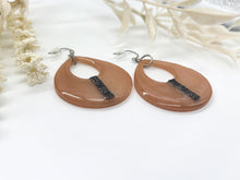 A pair of handmade peach-colored resin teardrop dangle earrings displayed from an angled, side-profile view on a clean white surface. The glossy, open cutout pendants showcase their thickness and a raised vertical stripe of glittering black rhinestones. They are fitted with silver-tone fishhook ear wires and clear rubber backs. Softly blurred white dried flowers frame the left and upper right sides of the composition.