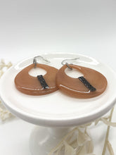A pair of handmade peach-colored resin teardrop dangle earrings displayed flat on a small, elevated white ceramic pedestal stand. The glossy open-cutout earrings feature a vertical stripe of glittering black rhinestones down the front. They are fitted with silver fishhook ear wires that rest towards the back. The background is a clean white, with soft, out-of-focus white floral elements visible below the stand.