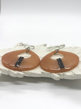 A low-angle shot of a pair of handmade peach-colored resin teardrop earrings showing  the thinness of them & how lightweight they are. They are resting flat on a textured, white ceramic feather plate. The glossy, open cutout earrings feature a vertical stripe of glittering black rhinestones aligned near the bottom. Silver-tone fishhook ear wires extend from the top of each piece. The foreground and background are clean and bright white, emphasizing the details and texture of the jewelry.