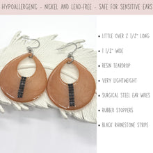 Product infographic for dangle earrings. Top banner reads: "Hypoallergenic - Nickel and lead-free - Safe for sensitive ears." The image shows a pair of peach-colored, cutout resin teardrop earrings with a vertical stripe of black rhinestones, resting on a white ceramic feather dish. Bullet points list details: a little over 2.5 inches long, 1.5 inches wide, very lightweight, surgical steel ear wires, rubber stoppers, and a black rhinestone stripe.