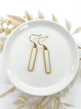 A pair of dangle earrings rests inside a small, glossy white ceramic dish. The earrings feature vertical, rectangular bar pendants filled with smooth white resin, framed by thin gold borders. The bars hang from gold fishhook ear wires. The dish is softly framed by blurred, light beige or cream-colored decorative leaves and dried florals in the background.