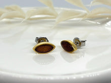 Close-up of handmade geometric marquise stud earrings displayed on a white ceramic surface. The earrings feature a gold-plated bezel filled with solid brown resin. One post is angled to display the side profile and surgical steel butterfly clutch, set against a soft-focus background.