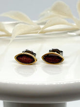 Pair of tiny minimalist geometric post earrings with a gold marquise bezel and a dark brown resin inlay, displayed on a glossy white surface with a soft-focus white leaf branch background.