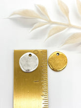Two metallic, organic round charms shown against a clean white background with a dried foliage accent. A silver charm rests atop a brass ruler to demonstrate size, measuring approximately 0.5 inches in diameter, while an identical gold charm lies adjacent to it. Both blank charms feature a top hole for hanging.