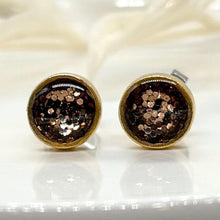 Close-up front view of handmade round stud earrings set in gold-plated bezels. The bezels feature a shimmering dark brown glitter resin inlay that catches the light. The pair rests on a smooth white surface with soft, out-of-focus botanical accents in the background.