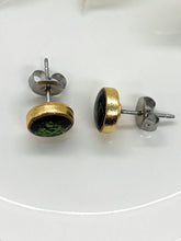 Angled side-profile view of handmade round stud earrings set in gold-plated bezels with emerald green glitter resin inlays. The left earring stands facing forward, showcasing its gold frame and surgical steel post with butterfly back. The right earring lays flat to highlight the side profile and hardware details against a clean white background.