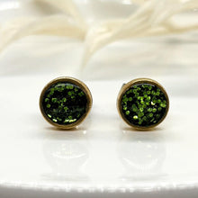 Close-up front view of handmade round stud earrings in gold-plated bezels. The bezels feature a vibrant green glitter inlay sealed in smooth resin that catches the light. The pair rests on a clean white surface with soft, out-of-focus white botanical accents in the background.