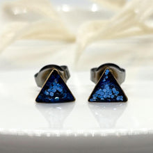 Handcrafted navy and bright blue glitter triangle stud earrings sealed in clear resin with surgical steel posts. Displayed on a white dish with neutral botanical accents. Modern geometric resin studs and boho gift for women by Jenica Jewelry.