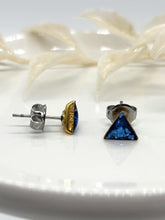 Side-profile view of blue glitter triangle stud earrings showing the gold bezel frame and hypoallergenic stainless steel post & backing. Displayed on a white ceramic dish with soft botanical accents in the background. Highlighting construction quality for sensitive ears by Jenica Jewelry.