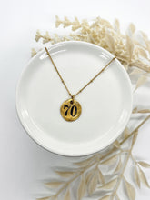 Handcrafted 70th birthday gold pebble charm necklace displayed flat on a round white dish. The organic round gold-plated pendant is hand-stamped with the number "70" in bold black ink, hanging from a delicate beaded satellite chain. Out-of-focus white dried florals frame the background for a soft, minimalist aesthetic.