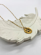 Angled close-up of a handcrafted gold pebble pendant necklace resting on a white textured ceramic feather dish. The organic round pendant is hand-stamped with the number "70" in black ink and attached to a delicate gold satellite chain, set against a clean white background.