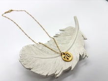 Full view of a gold hand-stamped pebble round charm necklace extended across a white ceramic feather dish. The organic round gold-plated pendant displays the number "70" stamped in black ink, attached to a dainty gold satellite chain against a bright, minimalist white background.